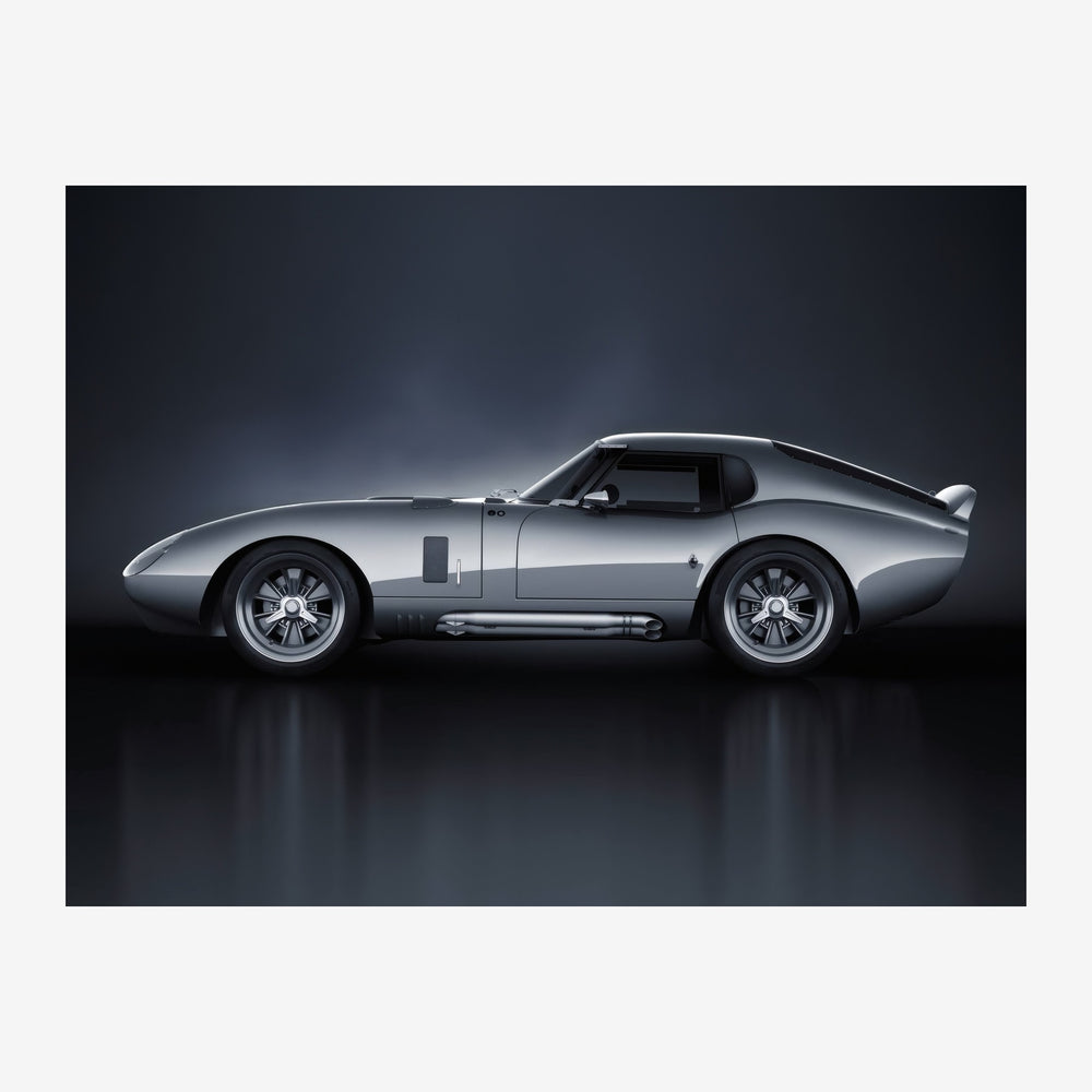 Voiture de sport classique argentée photographiée de profil sur un fond noir, avec son reflet visible au sol.