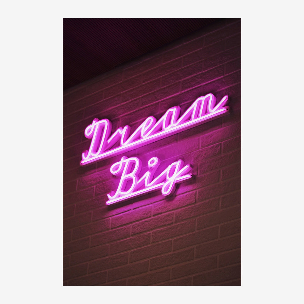 Enseigne lumineuse rose fluo « Dream Big » écrite en lettres cursives néon sur un mur de briques sombres.