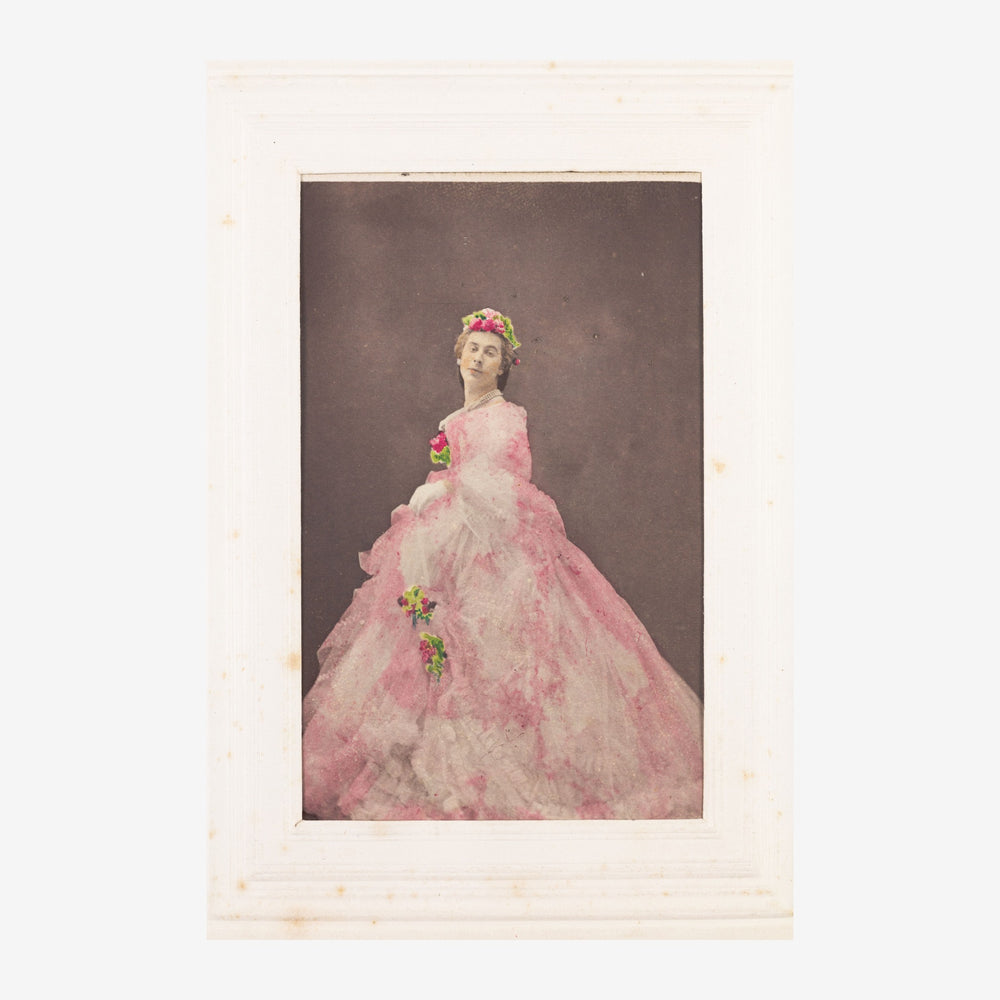 Portrait d'une femme portant une robe rose volumineuse avec couronne florale et fleurs à la main, sur fond taupe.