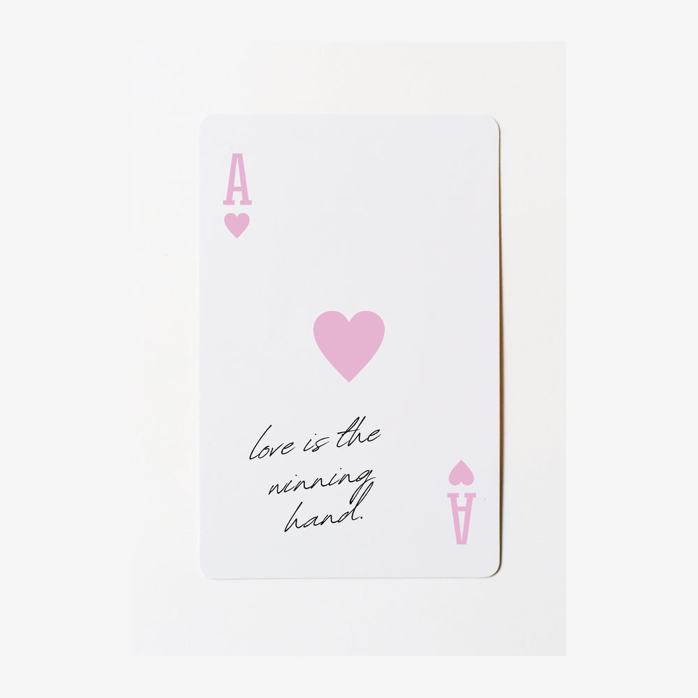Carte blanche ornée d'un cœur rose au centre, avec des symboles de cartes aux quatre coins et le texte manuscrit « love is.