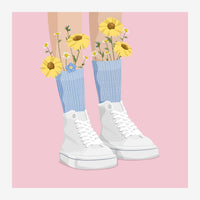 Deux baskets blanches à lacets remplies de marguerites jaunes et petites fleurs, sur fond rose poudré.
